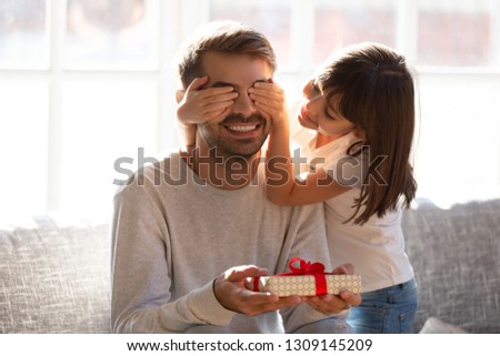 Cute kid daughter covering eyes of happy dad holding box sitting on sofa, little child girl making surprise preparing gift to smiling excited daddy on fathers day congratulating with birthday at home