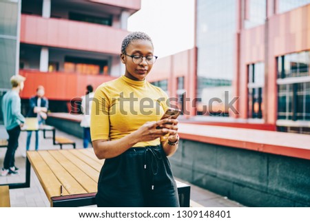 African american woman in eyeglasses holding modern smartphone and checking mail on device using 4G internet connection in urban setting.Dark skinned female reading received sms message on cellular