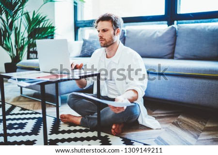 Skilled man concentrated on remote work and startup project sitting on floor in modern apartment and using 4g wireless for browsing online websites, young hipster guy typing text message via netbook