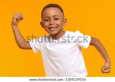 Body language. Picture of handsome Afro American 8 year old kid dressed in casual wear posing isolated at yellow wall, raising clenched fists and tensing bicep, demonstrating his strong arms