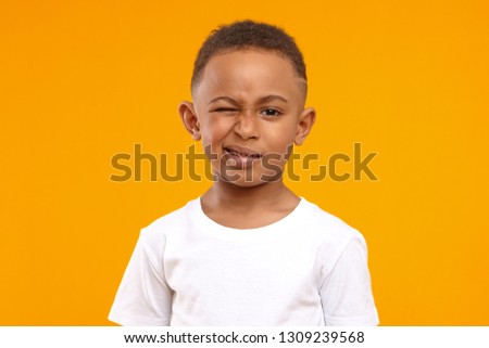 Children, fun and body language concept. Cute African American little boy grimacing against yellow studio wall background, winking at camera, planning joke or trick, wearing casual white t-shirt