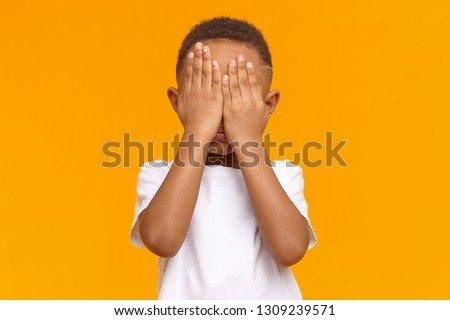 Shy little boy feeling ashamed because of his bad behavior, crying, not showing his tears. Isolated shot of unrecognizable male kid with hands on his face, being afraid of scary movie. Body language