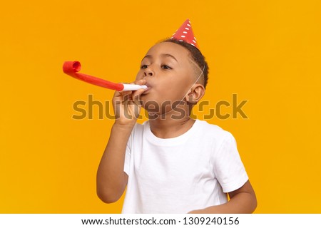 Happy Afro American little boy celebrating his ten year birthday with friends, posing isolated at blank yellow wall, blowing whistle, holiday hat on his head. Fun, joy, celebration and holiday