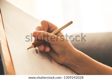 Close up view of a young hand drawing on a white sheet. Kid hold a black wooden pencil and draw something on a warm orange light at home. Children writing on a paper. Teen drawing freehand a manga.