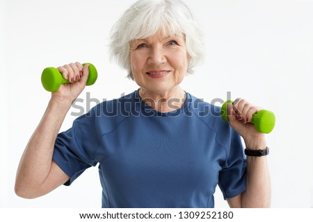 People, age, sports and active lifestyle concept. Picture of happy positive mature retired woman in t-shirt doing exercise with free weights in gym. Excited senior female training with dumbbells