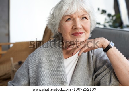 Elegance, age, beauty and people concept. Indoor image of charming elegant senior mature woman enjoying free time at home, sitting on couch in stylish living room interior, smiling happily Royalty-Free Stock Photo #1309252189