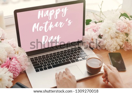 Happy Mother's Day text sign on pink laptop screen and girl hands with coffee, phone, black notebook and peonies on rustic wooden table. Stylish floral greeting card.