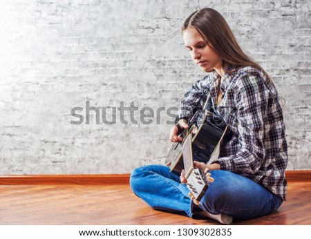 young teenager brunette girl with long hair sitting on the floor and playing an black acoustic guitar on gray wall background