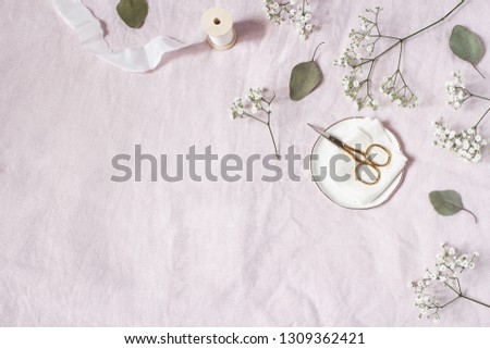 Styled stock photo. Feminine wedding desktop scene. Baby's breath Gypsophila flowers, dry green eucalyptus leaves, satin ribbon, golden scissors on pink linen background. Empty space. Top view. 