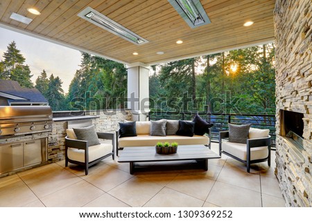 Luxury modern house exterior with covered patio boasting stone fireplace and cozy rattan furniture.
