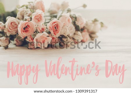 Happy Valentine's Day text sign on pink small roses on wooden background in light. Tender Flowers image. Valentines day floral greeting card.