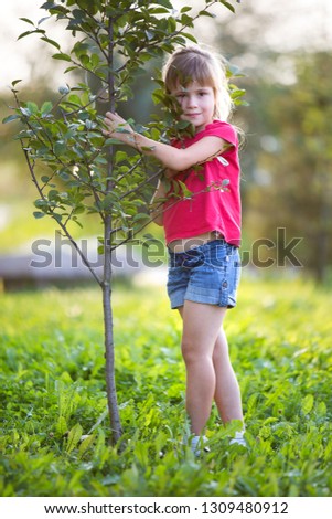 Cute pretty smiling child girl with gray eyes and long blond hair in summer clothing holding to young green sapling tree on blurred sunny park or garden bokeh background. Love to nature concept.