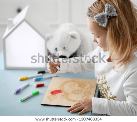 Kid girl in white shirt sits at the desk and paints hedgehog picture with multicolored markers accompanied by white bear