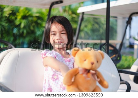 A lovely little girl sitting on a golf cart with her teddy bear. This portrait photo was taken in the park with soft morning light. Adorable kid concept.