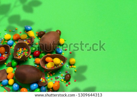 Broken and whole chocolate Easter eggs, multicolored sweets on a green background. Shrub. Concept of celebrating Easter, Easter decorations. Flat lay, top view. Copy Space.
