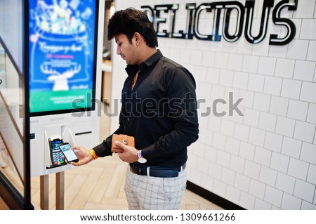 Indian man customer at store place orders and pay by contactless credit card on mobile phone through self pay floor kiosk for fast food, payment terminal. Pay pass.