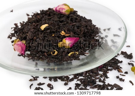 Dried leaves of black tea with chinese rose buds and petals of orange flowers on saucer and scattered beside to him close-up at selective focus on a white background

