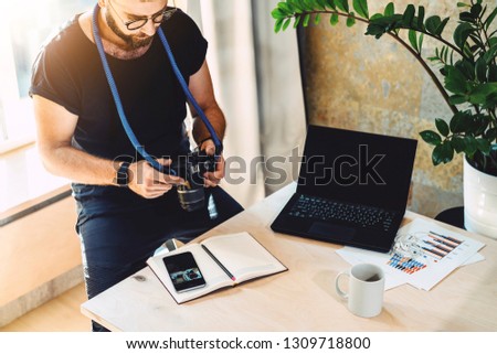 Man looks at camera screen,sitting in office at table with cup of coffee and laptop. Photographer is watching footage on camera screen. On desk notebook, smartphone.Concept of occupation and lifestyle