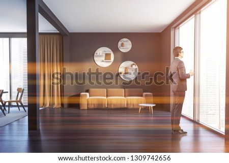 Man in interior of living room with gray walls, black wooden floor, panoramic window and beige sofa standing near round coffee table. Original round shelves above it. Toned image