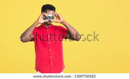 Young african american man holding a camera and photographing something on yellow background