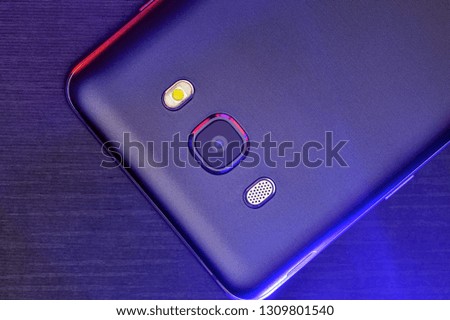 Black smartphone on dark background. A fragment of the cell phone case in black with red and blue backlight. The phone lies on the black technological ribbed surface of the camera up