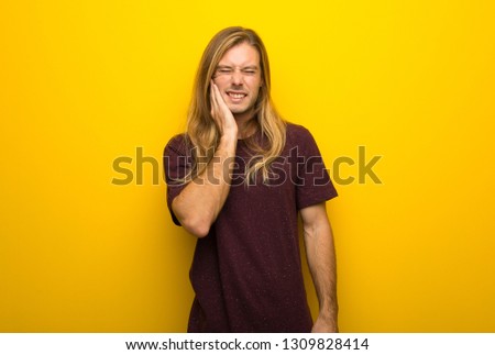 Blond man with long hair over yellow wall with toothache