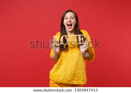 Funny young woman in yellow fur sweater keeping mouth wide open holding wooden word letters love isolated on bright red wall background. People sincere emotions, lifestyle concept. Mock up copy space