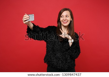Smiling young woman in black fur sweater pointing index finger doing selfie shot on mobile phone isolated on bright red wall background. People sincere emotions, lifestyle concept. Mock up copy space