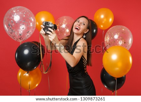 Cheerful young girl in black dress doing taking selfie shot on retro vintage photo camera on bright red background air balloons. Valentine's Day, Happy New Year, birthday mockup holiday party concept