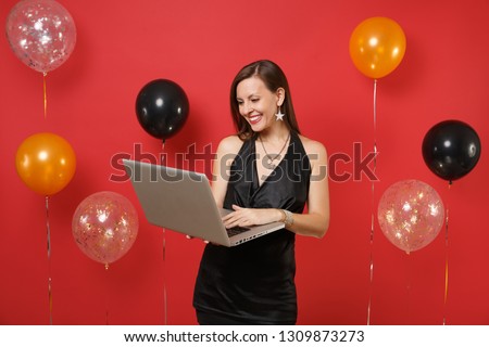 Pleasant young girl in little black dress working on laptop pc computer while celebrating on bright red background air balloons. Valentine's Day, Happy New Year birthday mockup holiday party concept