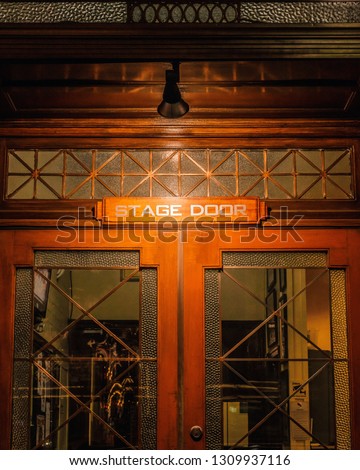 A 'Stage Door' sign with white lettering, on a wood and glass door with a lamp above