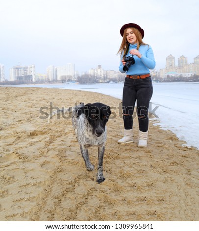 A young girl takes pictures of a dog on the beach near the river in the city. Winter landscape and people
