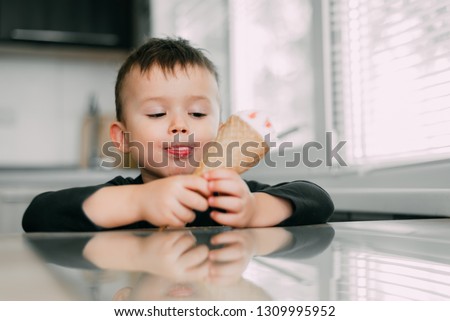 A child in a dark-blue t-shirt in the bright kitchen eating a waffle ice cream cone in the summer house