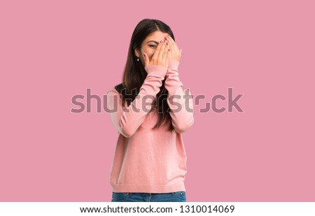 Teenager girl with pink shirt covering eyes by hands and looking through the fingers on isolated pink background