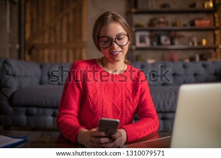 Beautiful smiling hipster girl in fashionable eye glasses reading article on blog via mobile phone, sitting with laptop computer in home during weekend. Cheerful woman using apps on cell telephone 