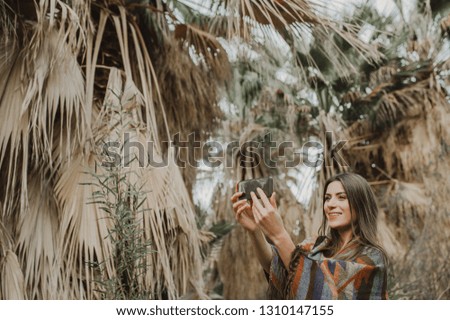 Smiling young woman holding smartphone, taking photos outdoors, surrounded by palms.