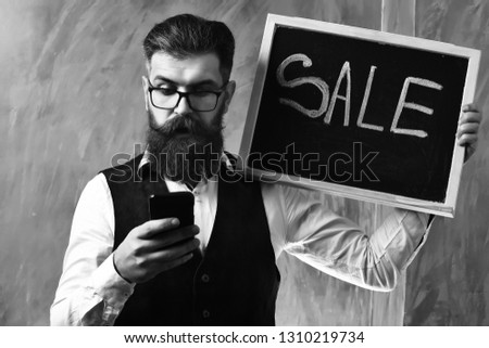 Bearded man, long beard. Brutal caucasian unshaven serious hipster with glasses and moustache surfing internet on cell phone, holding sale inscription on blackboard on brown studio background