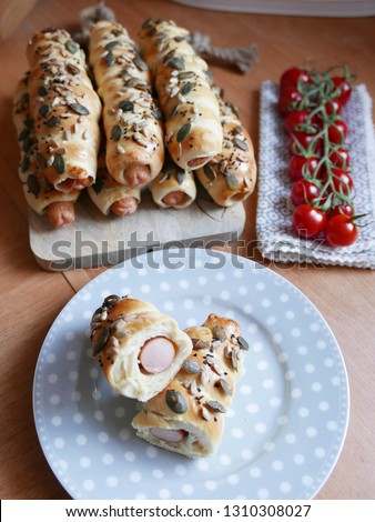 Hot Dogs wrapped in baked yeast dough with sunflower and pumpkin seeds, served on wooden cutting table and on plate with white dots, cocktail tomatoes in the background
