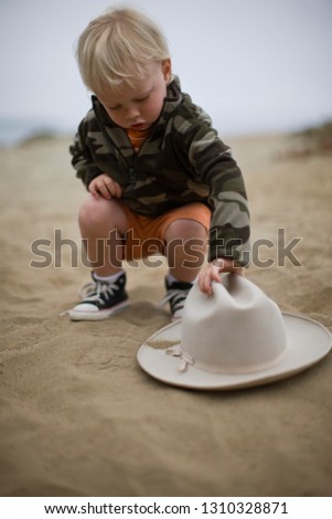 Curious toddler bending over to pick up a hat in the sand on a beach.