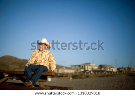 Smiling young boy wearing a cowboy hat while sitting on a picnic table on the beach in the sun.