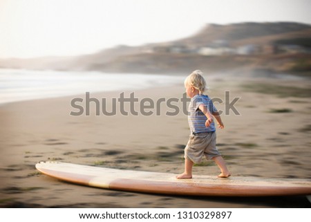 Young toddler standing on a surfboard resting on a sandy beach.