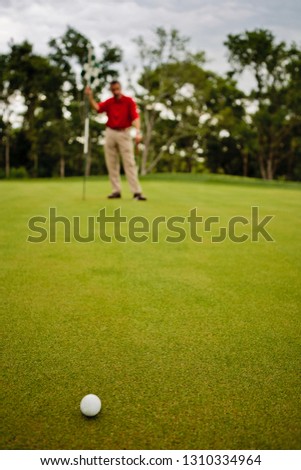 Mature man holds a golf flag in the background as a golf ball sits on the smooth grass in front of him.