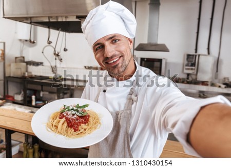 Image of happy man chief in apron taking selfie while standing at kitchen in restaurant