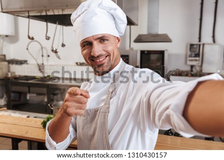 Photo of professional male chief in apron taking selfie while standing at kitchen in restaurant