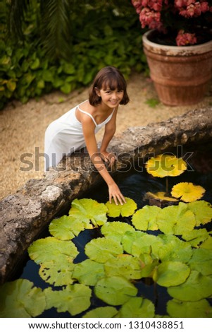 Portrait of a smiling young girl touching a lily pad in a lush garden fishpond.