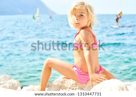 Child blond girl  sitting on the rock looking at camera with sea background. Summer vacation concept. 