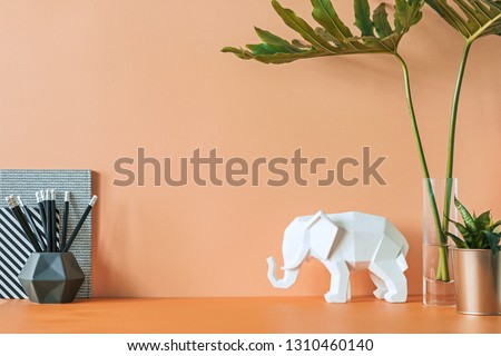 Orange interior of home office desk with leafs, elephant figure, plant and office accessories. Creative space with orange background wall. Creative workspace. Place for inscription.