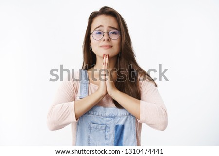 Waist-up shot of cute charming female brunette in glasses smiling and holding hands in pray raising eyebrows questioned as begging and waiting for positive answer while asking help over gray wall