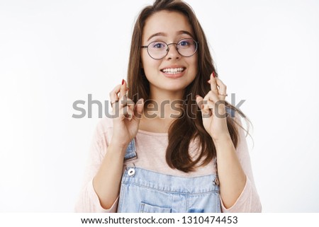 Girl crossing fingers with hopeful optimistic smile having faith dream come true, making wish to accomplish goals standing faithful with excited happy expression in glasses and denim overalls