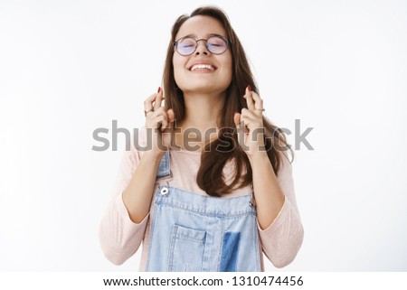 Waist-up shot of hopeful dreamy attractive woman in glasses raising head in sky with closed eyes and optimistic happy smile as praying begging god make dream come true, cross fingers for good luck